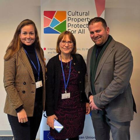 Three people (two women and one man) wearing conference badges and standing in front of a Cultural Property Protection for All banner