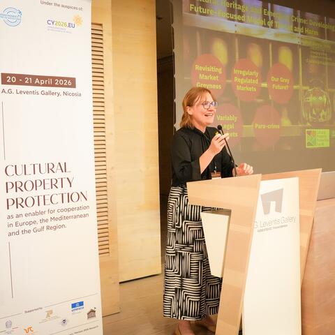 Woman standing at a conference podium speaking in front of a presentation screen.